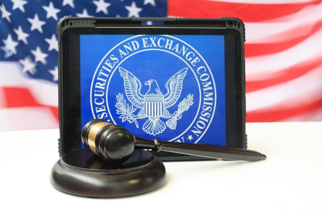 Ein Tablet auf dem das Logo der US Börsenaufsicht SEC abgebildet ist. Diese hat kürzlich ihre Klage gegen die Kryptowährung Ripple fallen lassen. Davor ist ein Richterhammer und im Hintergrund die US Flagge.